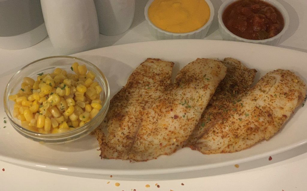 Picture of cooked tilapia on a oval white serving tray, flanked by corn. Queso and salsa dipping sauces frame the plate.